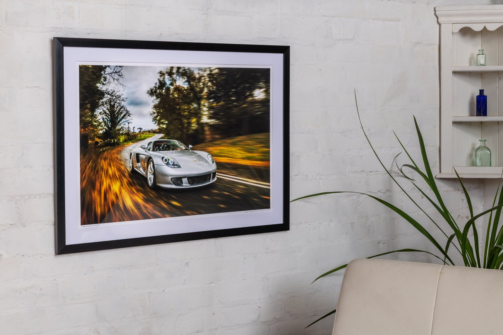 A framed print of a silver Porsche Carrera GT on an autumn road, hanging on a white brick wall